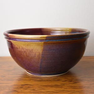 May include: A round ceramic bowl featuring a glossy glaze with blended tones of deep purple, dark blue, and warm golden yellow. The bowl has a smooth, curved silhouette with a slightly flared rim and a solid base, showcasing a variegated finish.