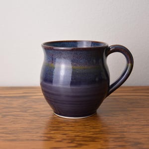 May include: A handmade ceramic mug with a dark blue and purple glaze. The mug has a rounded body, a curved handle, and a white base. It sits on a wooden surface.