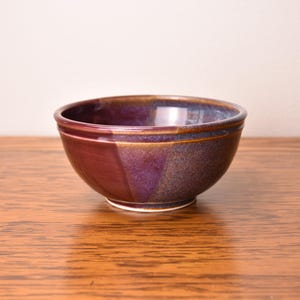 May include: A handmade ceramic bowl with a glossy finish. The bowl features a gradient of colors, including deep purple, burgundy, and touches of gold. The bowl is sitting on a wooden surface.