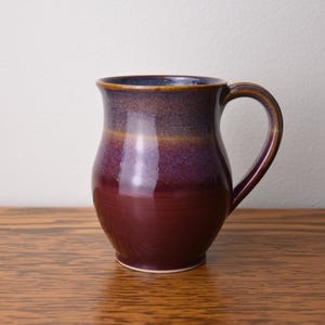 May include: A handmade ceramic mug with a glossy finish. The mug features a gradient of colors, transitioning from a deep burgundy at the base to purple and brown at the rim. It has a curved handle and sits on a wooden surface.