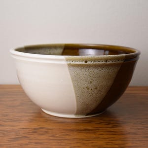 May include: A ceramic bowl featuring a two-tone design with a smooth, matte off-white finish on one side and a glossy, deep brown glaze on the other. A textured, speckled beige band separates the two colors, creating a distinct visual transition.