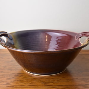 May include: A ceramic bowl with two side handles, featuring a deep burgundy and purple interior glaze that transitions into a dark brown exterior. The bowl has a smooth, glossy finish and a wide, open circular rim, showcasing a handcrafted design.