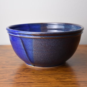 May include: A round ceramic bowl featuring a deep cobalt blue glaze that transitions into a darker, mottled navy and charcoal tone. The bowl has a smooth, curved silhouette with a distinct rim and a subtle, angled vertical line detail.