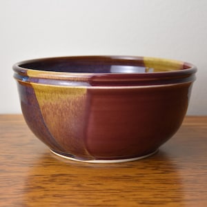 May include: A round ceramic bowl with a glossy finish, featuring a deep maroon and dark purple color palette with contrasting patches of mustard yellow glaze along the upper rim and side, showcasing a smooth, curved silhouette.