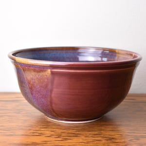 May include: A ceramic bowl with a deep burgundy exterior and a glossy finish. The rim has a band of blue and gold. The bowl sits on a wooden surface.