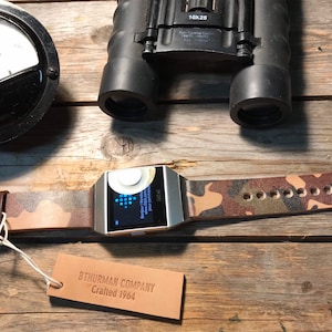 Puede incluir: Un reloj inteligente plateado con una correa de cuero con estampado de camuflaje. La esfera del reloj muestra un reloj digital azul y la palabra "Fitbit". Una etiqueta de cuero marrón con el texto "BThurman Company Crafted 1964" está en primer plano.