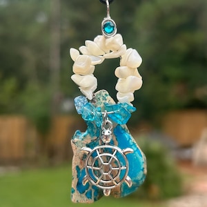 May include: A handcrafted pendant necklace featuring a silver turtle charm, white shell accents, and blue gemstone chips. The pendant hangs from a black cord and includes a teal-colored gemstone at the top. The pendant is approximately 2 inches long.