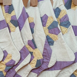 May include: A set of six white fabric stockings with purple and yellow patchwork designs. The stockings are hanging from a wooden surface.