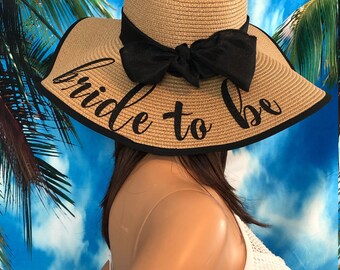 May include: A wide-brimmed straw hat with a black ribbon bow. The hat has a black trim and the words "bride to be" are written in black script across the brim. The hat is a beach or bridal accessory.