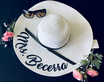 May include: White sun hat with a black ribbon and the text "Mrs. Becerra" embroidered on the brim. Sunglasses are on top of the hat.
