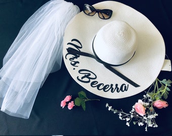 May include: White sun hat with a black ribbon and the text "Mrs. Becerra" embroidered on the brim. The hat is surrounded by white flowers and a white veil.