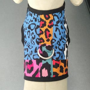 May include: A colorful dog harness with a patchwork design. The harness features a mix of patterns, including blue leopard print, orange and red leopard print, and pink and black leopard print. It has a black trim and a silver metal D-ring.
