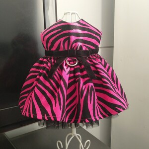 以下が含まれることがあります： ピンクと黒のゼブラ柄のドレス。黒いベルベットのベルトとシルバーのバックルが付いています。ドレスはフレアスカートと黒のチュール裾が特徴です。白いマネキンに飾られています。