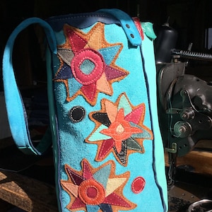 May include: A turquoise blue leather bag with a long strap. The bag is decorated with three colorful star-shaped appliques made of leather scraps. The appliques are in shades of red, orange, yellow, green, blue, and purple.