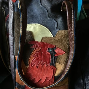 May include: A brown leather tote bag with a red cardinal applique. The bag is made of different pieces of leather sewn together, creating a patchwork design. The cardinal is stitched with red thread and has a blue eye.