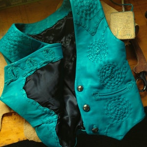 May include: A teal leather vest with intricate stitching and button closures. The vest has a black lining and is made of a soft, supple leather.