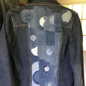 May include: A blue denim jacket with a patchwork design on the back. The patchwork is made of different shades of blue denim and is arranged in a geometric pattern.