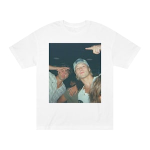 Puede incluir: Camiseta blanca con una fotografía de tres jóvenes. Uno lleva una gorra de béisbol y señala. Otro hace un gesto de 'ok'. La imagen tiene un aspecto vintage, ligeramente descolorido.