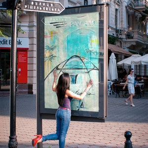 May include: A woman in blue jeans and a red shirt poses in front of a billboard with a blue and white abstract design. The billboard has the text "PAN AM" and "вулиця ришельєвська" on it.