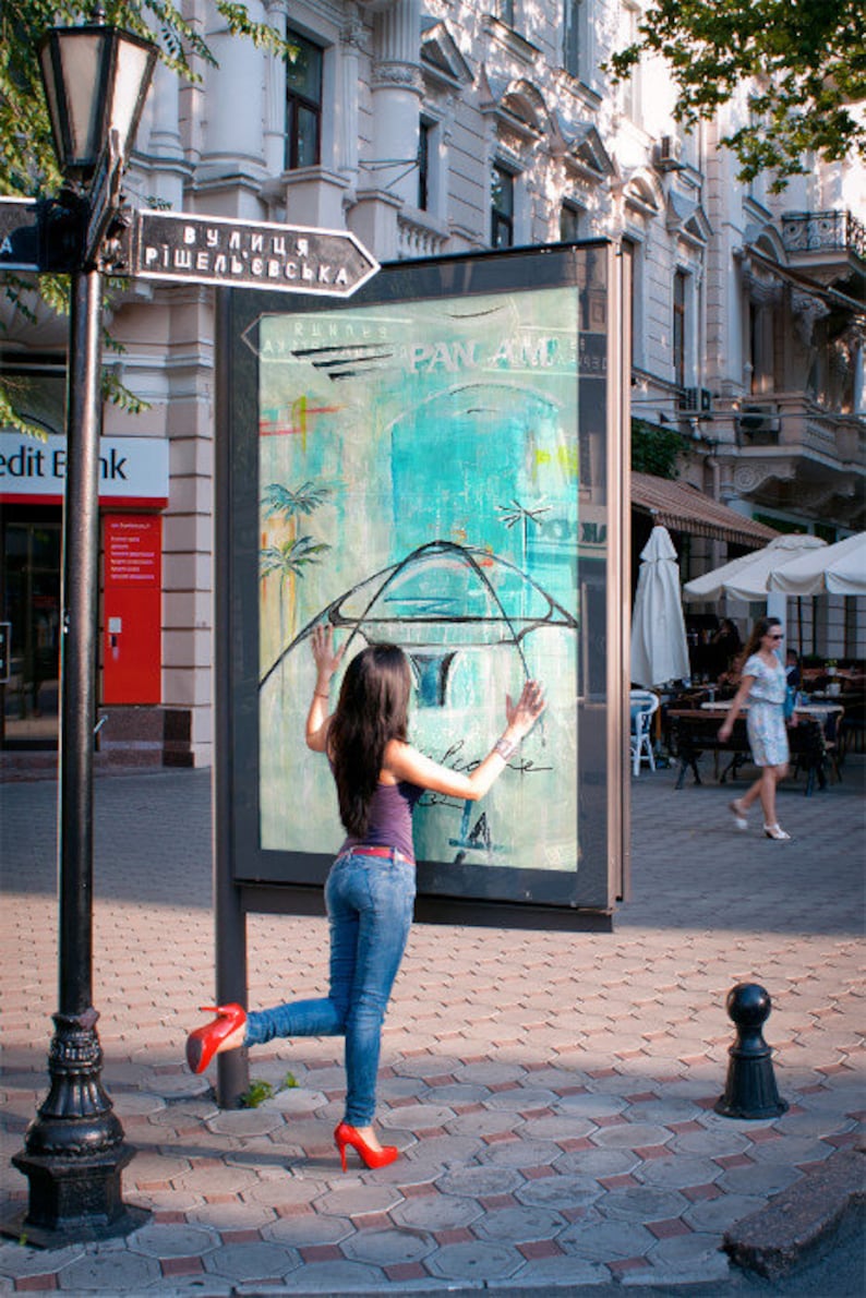 May include: A woman in blue jeans and a red shirt poses in front of a billboard with a blue and white abstract design. The billboard has the text "PAN AM" and "вулиця ришельєвська" on it.