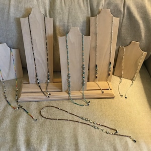 May include: A collection of seven beaded eyeglass chains displayed on wooden stands. The chains are made with various colors and patterns of beads, including black, brown, white, blue, green, and red.