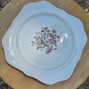May include: A white ceramic plate with a scalloped edge and a gold-trimmed rim. The center of the plate features a detailed floral bouquet illustration containing pink, yellow, and green flowers with dark green leaves on a smooth white surface.
