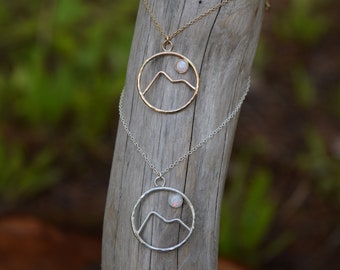 Puede incluir: Dos collares con colgantes de montaña y sol. Uno es dorado, el otro plateado. Cada colgante presenta un marco circular con una silueta de montaña y una pequeña piedra de ópalo redonda que representa el sol.