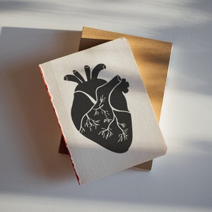 May include: A black and white illustration of a human heart on a white notebook with a brown notebook underneath.
