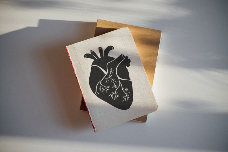 May include: A black and white illustration of a human heart on a white notebook with a brown notebook underneath.