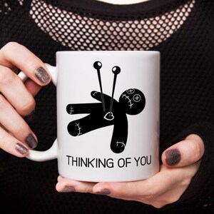 May include: White ceramic mug with a black and white illustration of a voodoo doll with needles sticking out of it. The text "Thinking of You" is printed below the illustration.