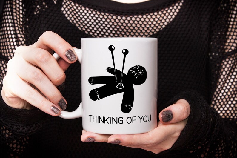 May include: White ceramic mug with a black and white illustration of a voodoo doll with needles sticking out of it. The text "Thinking of You" is printed below the illustration.