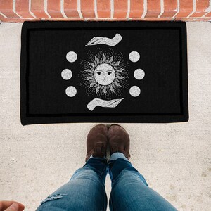 May include: Black doormat with a white sun and moon design. The sun has a face and is surrounded by six moons. The design is framed by two hands.