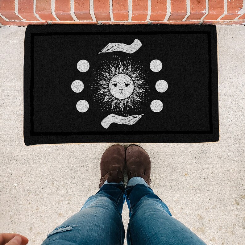 May include: Black doormat with a white sun and moon design. The sun has a face and is surrounded by six moons. The design is framed by two hands.
