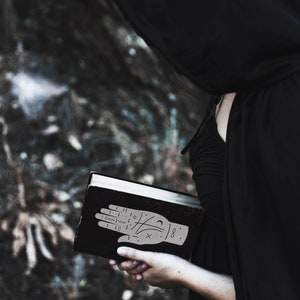 May include: A person wearing a black hooded cloak holds a black book with a hand drawn on the cover. The hand has lines and symbols drawn on it, and the number 8 is written on the wrist.
