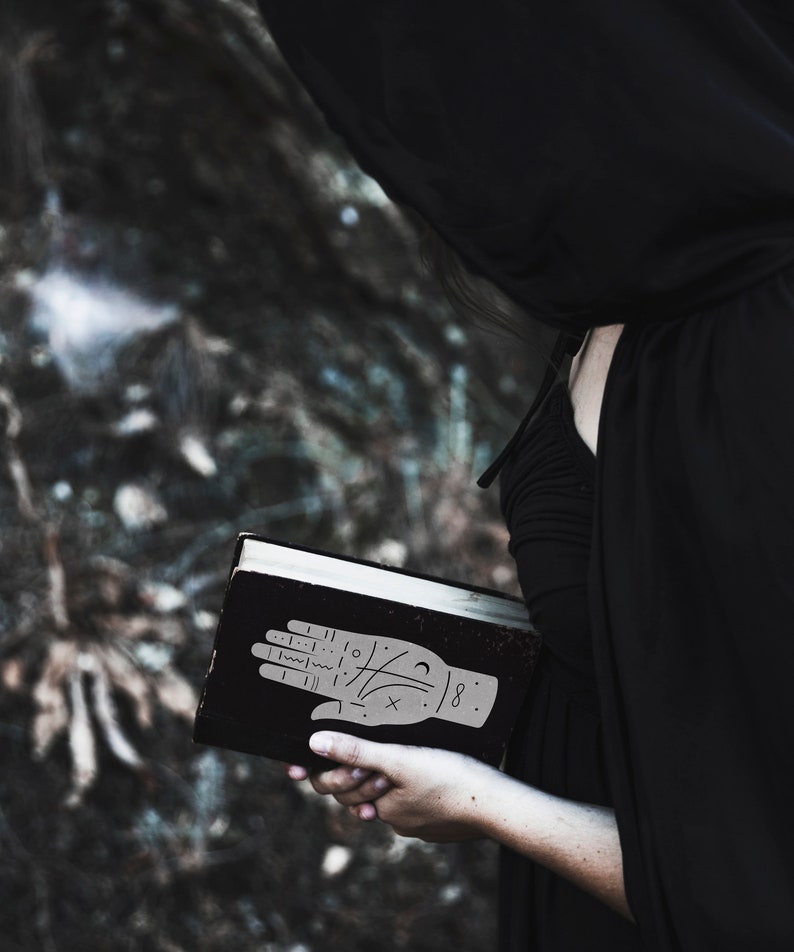 May include: A person wearing a black hooded cloak holds a black book with a hand drawn on the cover. The hand has lines and symbols drawn on it, and the number 8 is written on the wrist.