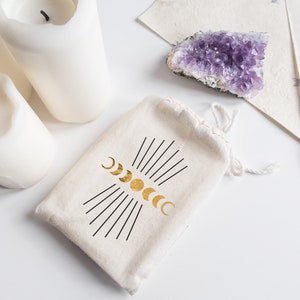 May include: A white cotton drawstring bag with a gold moon phases design. The bag is sitting on a white surface with three white candles and a purple amethyst crystal.