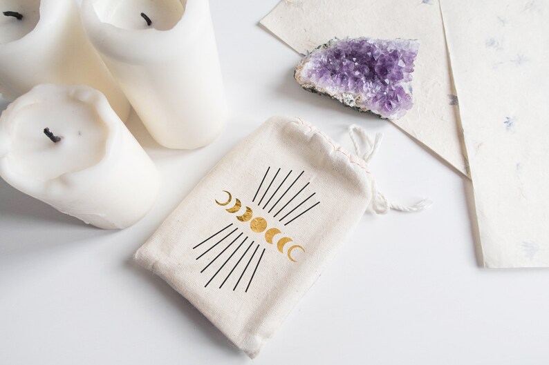 May include: A white cotton drawstring bag with a gold moon phases design. The bag is sitting on a white surface with three white candles and a purple amethyst crystal.