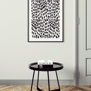 Puede incluir: Una impresi&oacute;n abstracta en blanco y negro en un marco negro cuelga de una pared blanca sobre una mesa auxiliar redonda negra con una tapa de vidrio negro. Un libro titulado "Vogue" est&aacute; encima de la mesa.