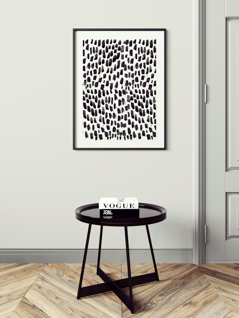 Puede incluir: Una impresi&oacute;n abstracta en blanco y negro en un marco negro cuelga de una pared blanca sobre una mesa auxiliar redonda negra con una tapa de vidrio negro. Un libro titulado "Vogue" est&aacute; encima de la mesa.
