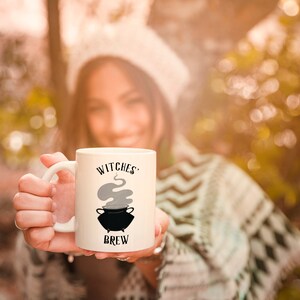 Może przedstawiać: Biały ceramiczny kubek z czarno-szarym rysunkiem kotła z unoszącą się z niego parą. Na kubku nadrukowany jest napis "Witches' Brew".