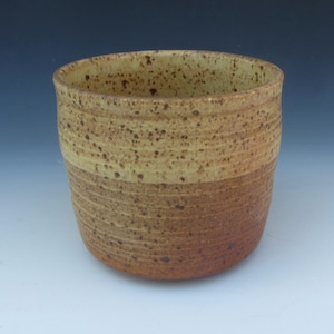 May include: A handmade ceramic tumbler with a speckled, textured surface. The upper half is a light tan color, while the lower half transitions to a darker brown. The cup has a cylindrical shape and is suitable for beverages.