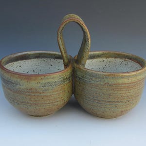 May include: A double ceramic bowl set with a central arched handle. The exterior features a mottled, earthy brown and green glaze with horizontal striations, while the interior is a speckled off-white color with a smooth, rounded rim design.