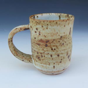 May include: A set of three ceramic mugs with a speckled, cream-colored glaze. Each mug features a different hand-painted design: a blue and green leaf, a yellow flower, and a red berry. The mugs have a simple, rounded shape.