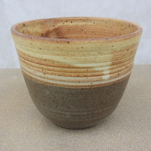May include: A handmade ceramic bowl with a wide, open top. The bowl features a two-tone design, with a dark brown base and a lighter tan upper section. The upper section has horizontal stripes of tan and cream colors. The bowl is suitable for serving or display.