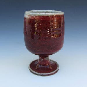 May include: A ceramic goblet featuring a deep red, mottled glaze with subtle variations in tone. The vessel has a rounded bowl, a short, thick stem, and a circular base, with a light-colored, unglazed rim that contrasts with the dark red finish.