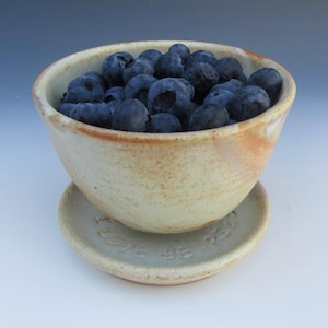 May include: A speckled beige ceramic bowl filled with fresh blueberries, resting on a matching circular saucer. The saucer features the embossed text I LOVE BLUEBERRIES. The bowl has a rounded shape with subtle orange-toned glaze accents.