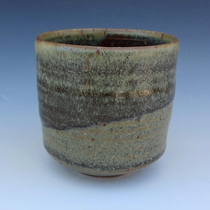 May include: A cylindrical ceramic cup featuring a mottled glaze with layers of dark brown and earthy beige tones. The surface has a speckled, textured appearance with a smooth rim and a rounded base, showcasing a blend of dark and light colors.