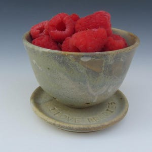 May include: A speckled, light-toned ceramic berry bowl with drainage holes, filled with fresh red raspberries. The matching saucer base features the embossed text I LOVE BERRIES. The bowl has a rustic, earthy finish with subtle grey and tan tones.