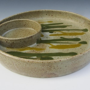May include: A speckled ceramic serving dish with a built-in dip bowl. The dish is beige with green and yellow paint streaks. The dip bowl is a smaller, circular indentation within the larger dish.