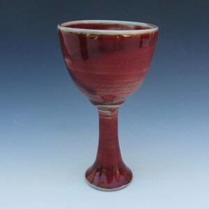 May include: A ceramic goblet featuring a deep red glossy glaze with subtle variations in tone. The vessel has a wide, rounded bowl that tapers into a slender, elongated stem, ending in a flared circular base with a visible white rim edge.